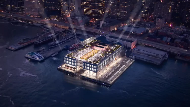 Aerial night view of The Beekman hotel on a pier, lit up with rooftop greenery, boats, and city skyline in background.