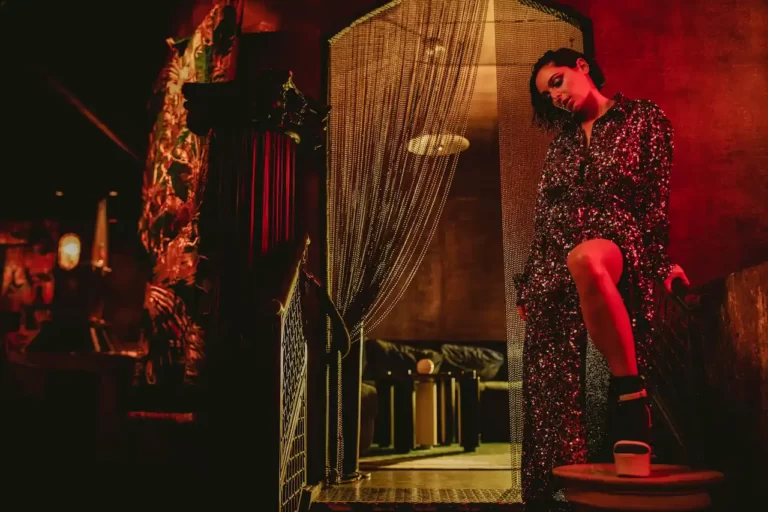 Woman in sparkling dress poses confidently under dramatic lighting, standing near rope curtain in stylish lounge.