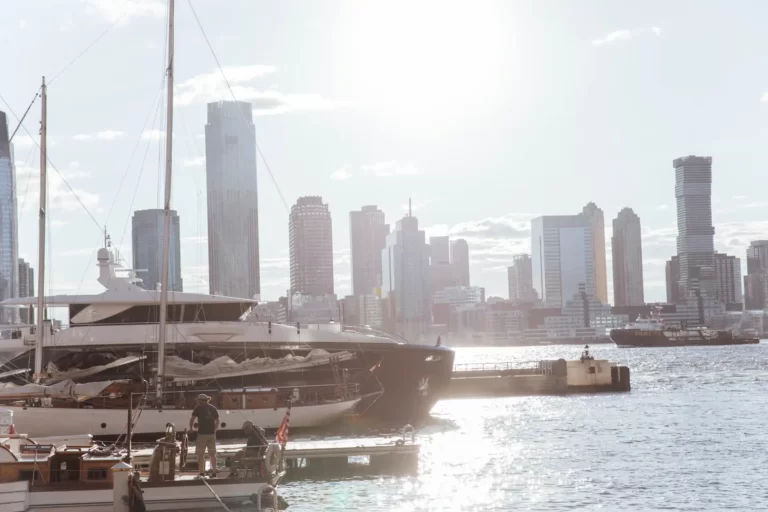 Large luxury yacht docked on city waterfront, with bright sunlight and modern skyscrapers in the background.