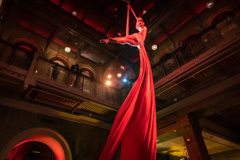 Aerial silk performer suspended mid-air in red silks, illuminated inside an ornate venue with arched windows.