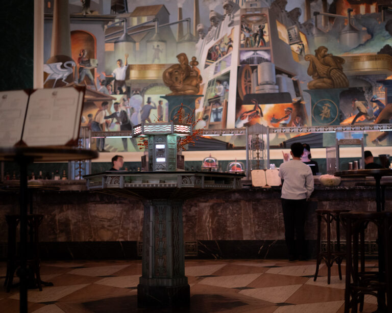 Interior of a bar with geometric tile flooring and a large mural depicting workers, machinery, and mythological figures.