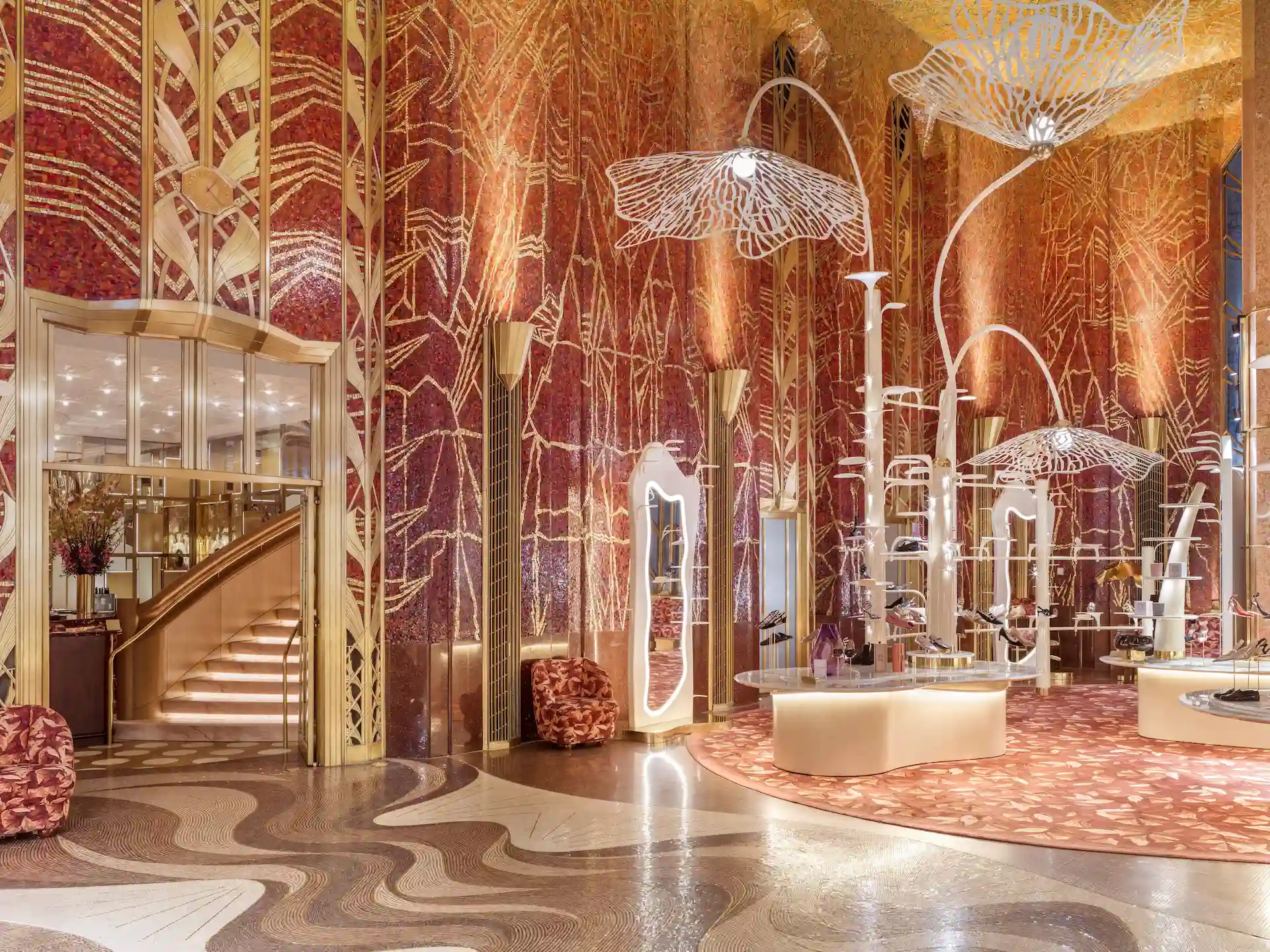 Art Deco–style lobby with red‑and‑gold mosaic walls, sculptural light fixtures, patterned floor, and a lit grand staircase.